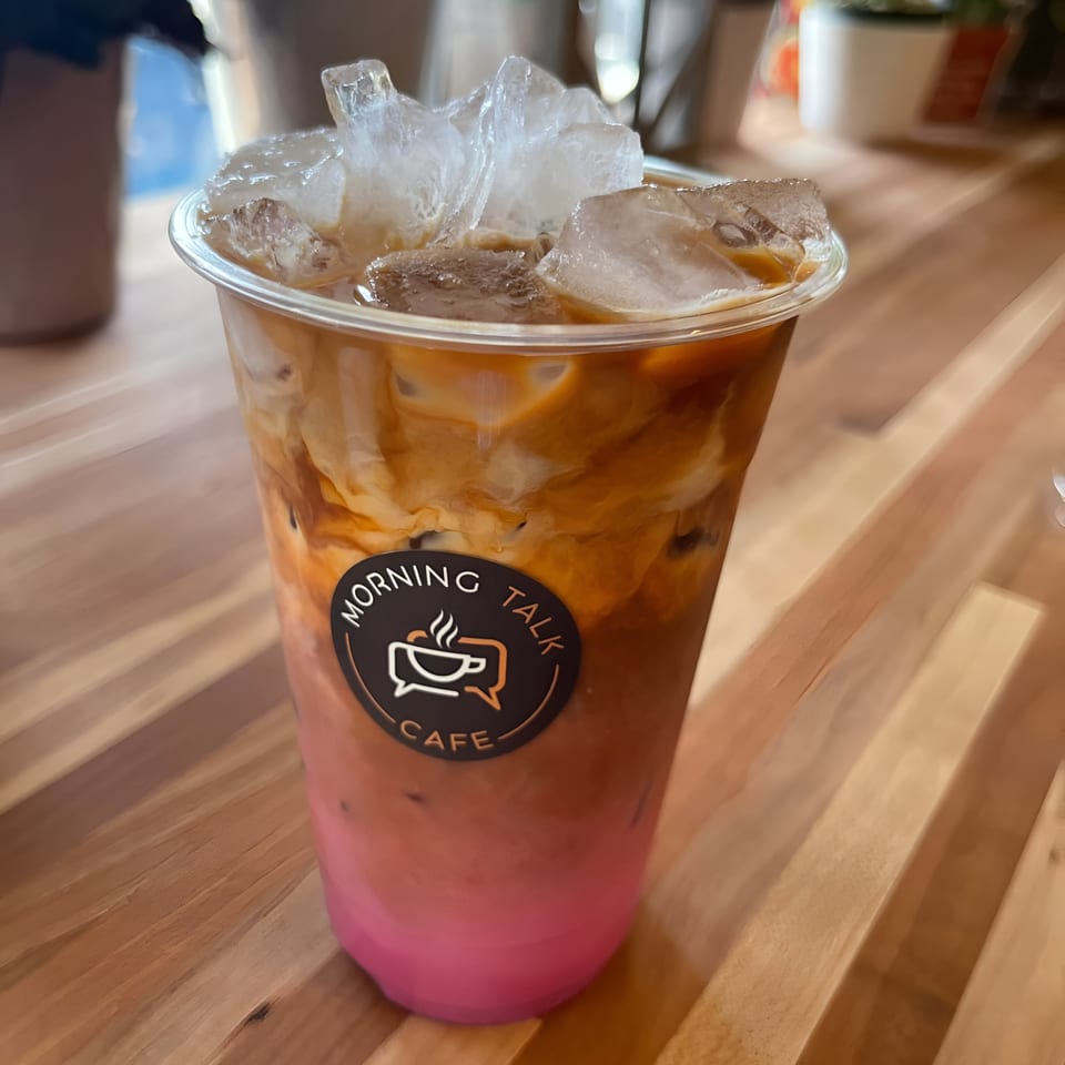Iced coffee drink with pink and brown layers, Morning Talk Cafe logo, on a wooden table.