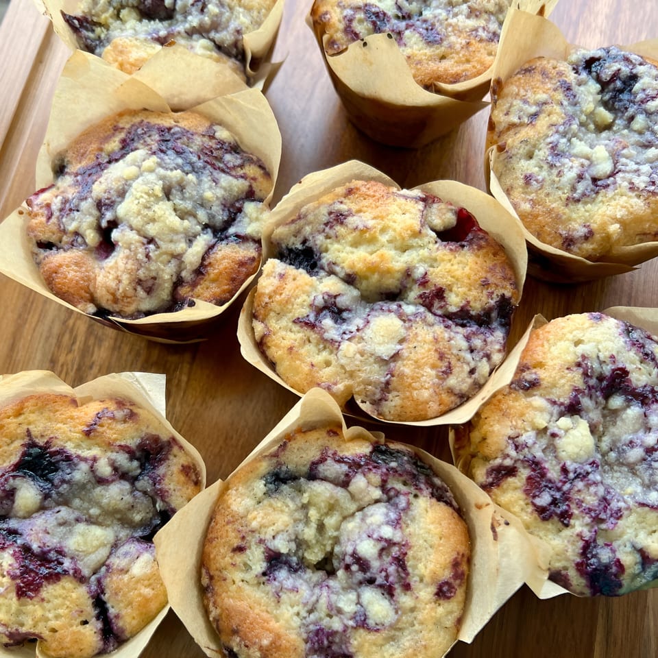 Several blueberry muffins with crumb topping in paper liners on a wooden surface.