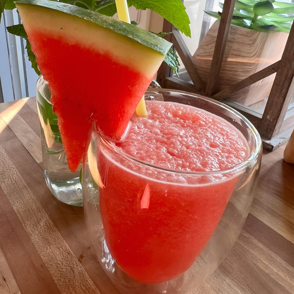A Morning Talk Cafe watermelon smoothie with a watermelon slice and straw, on a wooden table.