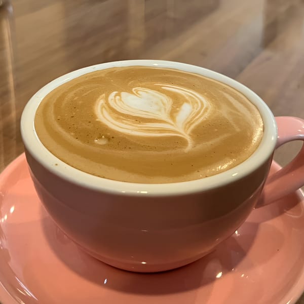 A pink cup of latte with a heart-shaped foam design sits on a matching pink saucer.